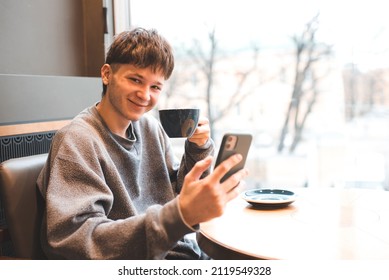 Smiling Teenage Gen Z Boy 17-18 Year Old Drinking Coffee And Holding Mobile Phone Sitting In Cafe. Happy Millennial Man Chat With Friends With Cup Of Tea Have Good Time. Working Place.