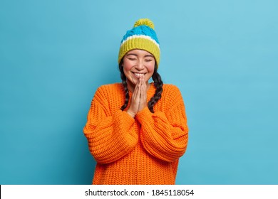 Smiling teenage girl enjoys winter time keeps palms together and laughs happily with closed eyes being amused by friend dressed in knitwear isolated on blue background. Positive emotions concept
