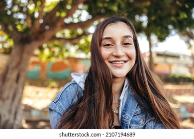 Smiling teenage girl looking at the camera outdoors. Happy young teenager wearing a denim jacket in the city. Female youngster sitting alone in an urban park during the day. - Shutterstock ID 2107194689