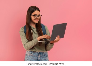 Smiling teenage girl wearing eyeglasses and backpack looking at laptop screen, cute female student symbolizing education and online learning, standing against minimalistic pink background, free space - Powered by Shutterstock