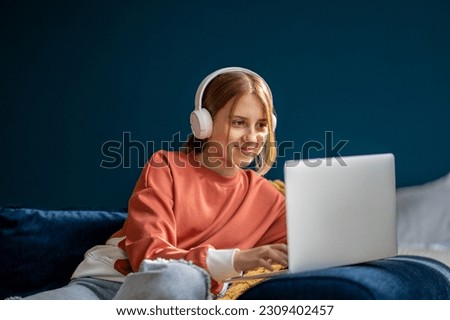 Smiling teenage girl wearing headphones typing on laptop, listening music and chatting online with friends. Happy teen using computer during leisure time, child teenager enjoying distance learning