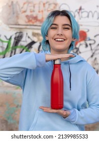 Smiling Teenager In Blue Hoodie With Red Water Bottle. Blue Haired Teen Girl Staying Outdoors Near Graffiti Wall. Reusable Bottle Mockup.  Hipster And Adolescence Concept