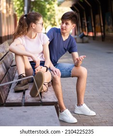 Smiling teenager boy talking with cute girl sitting on bench on city street on sunny summer day