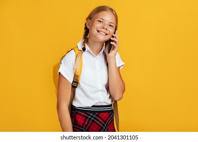 Smiling Teenager Caucasian Blonde Female With Pigtails, Backpack Speaks By Phone, Isolated On Yellow Background, Copy Space. Gadget, Parental Control, Be Safe, Back To School And Pupil Communication