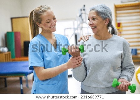 Smiling therapist assisting senior woman exercising with dumbbells at rehabilitation center