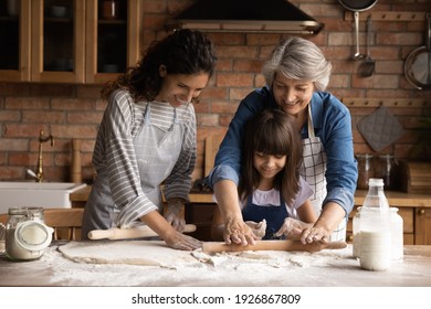 Smiling three generations of Hispanic women have fun baking together with dough at home kitchen. Happy little Latino girl child with young mom and senior grandmother cook pastries or cookies. - Powered by Shutterstock