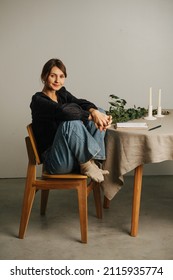 Smiling Tired Woman Sitting With Legs Up On A Chair Next To A Round Table. She's Wearing Mom Jeans Over Black Dress. Inside A Room