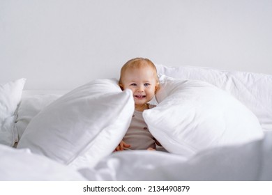 Smiling Toddler Having Fun On Bed With Lots Of Pillows. A Baby Playing Hide And Seek Or Peek A Boo.