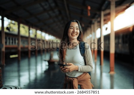 Smiling tourist with a backpack, checking her mobile phone while waiting for a train at Bangkok Hua Lamphong railway station, enjoying the vibrant atmosphere of Thailand