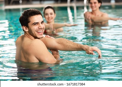 Smiling trainer looking at camera while exercising with young people in swimming pool