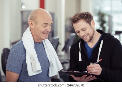 Smiling Trainer Working With A Senior Man At The Gym Writing Notes On A Clipboard With A Smile Of Encouragement In A Health And Fitness Concept