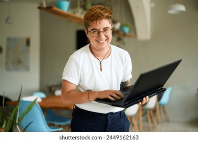 Smiling transgender professional holds laptop, works happily in modern office space. Inclusive workplace, young trans person enjoys career, experiences job fulfillment with tech in eco-friendly area.