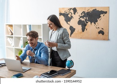 Smiling Travel Agent Pointing With Hand At Computer Monitor Near Attractive Colleague Holding Cup Of Coffee