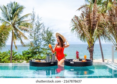 Smiling Travel Woman Or Influencer Drinking Orange Juice In Infinity Transparent Swimming Pool In Luxury Hotel. Exotic Summer Vacation On Beach. Tropical Sea Holidays