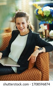 Smiling Trendy 40 Years Old Woman In White Blouse And Black Jacket With Closed Laptop Sitting On Couch In The Modern Living Room In Sunny Day.