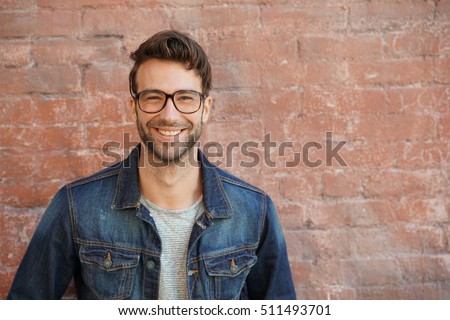 Smiling trendy guy with blue jeans jacket and eyeglasses standing by brick wall