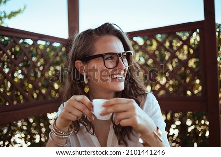 smiling trendy middle aged housewife in white shirt with eyeglasses drinking coffee in the patio of guest house hotel.