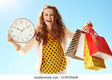 Smiling Trendy Woman In A Light Jacket Showing Shopping Bags And Clock Against Blue Sky