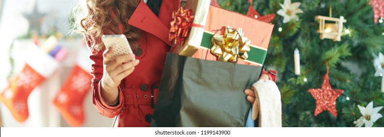 smiling trendy woman in red trench coat with shopping bag full of Christmas present boxes sending text message from phone near Christmas tree