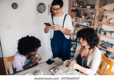 Smiling tutor standing at ceramics workshop and taking pictured of multicultural pottery class students doing pottery. Portrait of happy female instructor taking pictures with phone at pottery studio. - Powered by Shutterstock