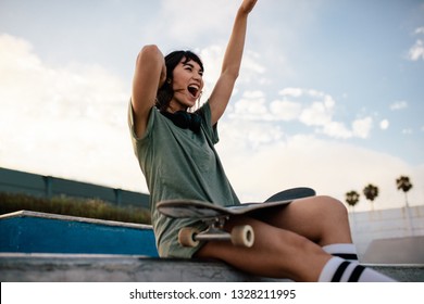 Smiling Urban Girl Sitting On Skateboard Ramp In Skate Park Having Fun. Female Skateboarder Enjoying Herself Outdoors At Skate Park.