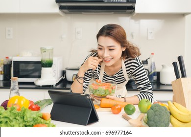 Smiling Vietnamese Woman Watching Tv Show On The Digital Tablet And Cooking Dinner In Home Kitchen