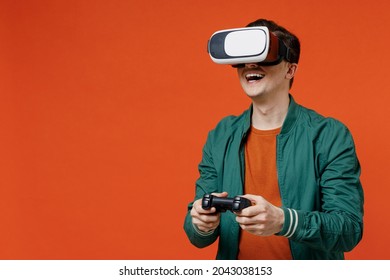 Smiling vivid excited happy young brunet man 20s wear red t-shirt green jacket watching in vr headset pc gadget play pc game with joystick console isolated on plain orange background studio portrait.