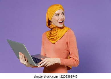 Smiling Vivid Young Arabian Asian Muslim Woman In Abaya Hijab Yellow Clothes Hold Use Work On Laptop Pc Computer Looking Back Behind Isolated On Plain Pastel Light Violet Background Studio Portrait