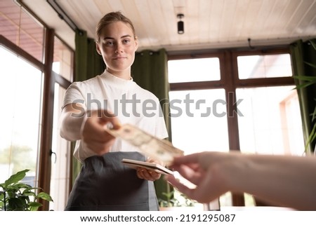 smiling waitress in black apron ready to accept visitor's money, attractive young woman taking dollars, looking at client, Concepts for payment, service charge, bill checking, money tips.