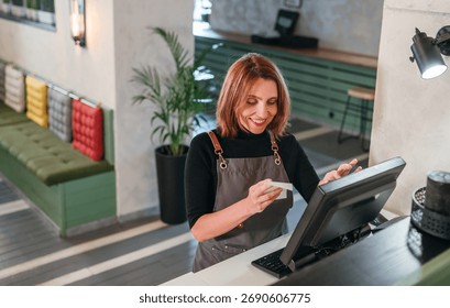 Smiling waitress in black uniform using point-of-sale terminal to process customer orders. Bartender at work in restaurant, hospitality service, and gender equality in the food and beverage industry.