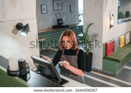 Smiling waitress in black uniform using point-of-sale terminal to process customer orders. Bartender at work in restaurant, hospitality service, and gender equality in the food and beverage industry.