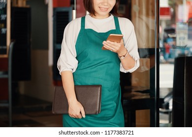 Smiling waitress with menu in her hands texting friend during short break