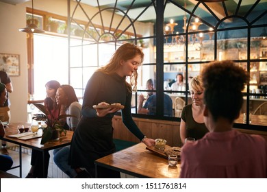 Smiling waitress serving an order of food to a group of young friends having drinks and eating together in a bistro