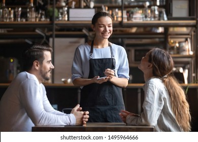 Smiling waitress wear apron hold notepad pen take order talk to clients serving restaurant guests couple choosing food drinks menu sit at cafe coffeehouse table, waiting staff, good customer service - Shutterstock ID 1495964666