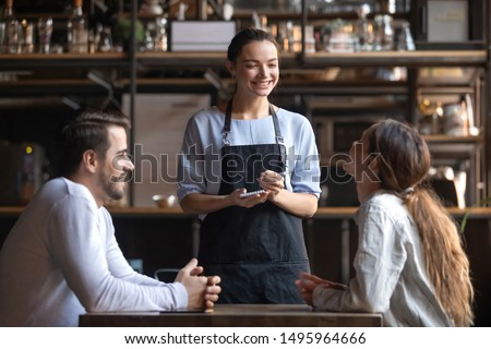 Smiling waitress wear apron hold notepad pen take order talk to clients serving restaurant guests couple choosing food drinks menu sit at cafe coffeehouse table, waiting staff, good customer service