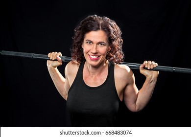 A Smiling Weight Trainer Poses For The Camera With A Barbell On Her Back.  She Is On A Black Background And Is Cross Lit.