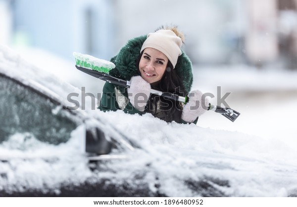 Smiling well clothed\
woman holds ice scraper and snow broom while leaning against the\
car covered in snow.