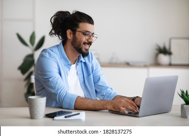Smiling western freelancer guy working on laptop at home office, sitting at desk using computer, young indian man in eyeglasses typing on keyboard, looking at screen, enjoying remote work, copy space - Shutterstock ID 1828137275