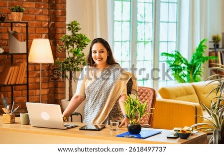 Smiling while showing thumbs up gesture, Indian young businesswoman or corporate female employer in ethnic saree confidently standing beside laptop at office desk looking at camera in cozy workspace