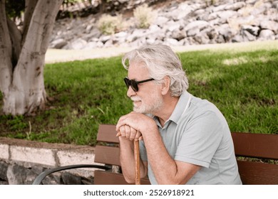Smiling white haired elder 70s man with walking disability holding knob of cane,sitting  in a bench in the park, suffering from muscles problems, bones disease, recovering after trauma