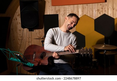 Smiling white man tuning guitar, playing, holding musical instrument in hands, sitting on chair in studio. Darken photo. Male wears casual cloth. Hobby, leisure of creative person, artist.Horizontal