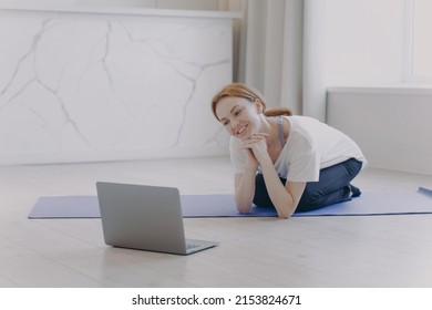 Smiling White Woman Has Remote Personal Training And Talking In Front Of Computer. Young Pretty Sportswoman Sitting Kneeling On Floor On Mat. Concept Of E-learning. Sport While Coronavirus Quarantine.