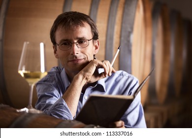 Smiling winemaker in cellar looking satisfied at a glass of white wine during wine tasting, with notebook and pen in hands.