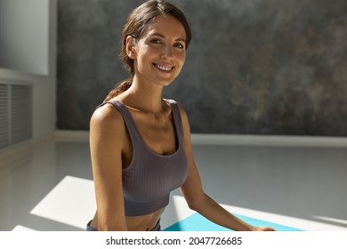 Smiling woman of 30's looking at camera practicing yoga, sitting in Padmasana, dressed in lilac stylish fitness bra, having long brown curly hair tied in ponytail. Wellness, healthy lifestyle - Powered by Shutterstock