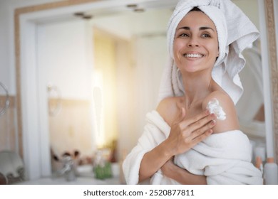 Smiling woman applies lotion to her shoulder, wrapped in towel, in bright bathroom. Her towel-wrapped hair and white bathrobe suggest self-care or skincare routine, conveying wellness and relaxation - Powered by Shutterstock