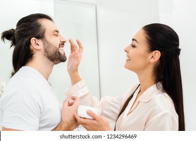 Smiling Woman Applying Face Cream On Man Nose In Bathroom