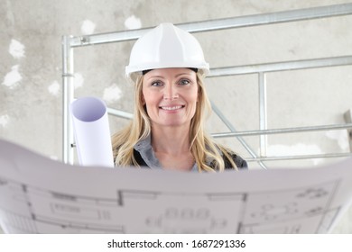 Smiling Woman Architect Or Construction Engineer Reading Blueprint Wear Helmet Inside A Building Site With Scaffolding In The Background