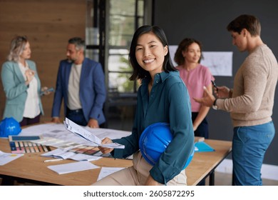 Smiling woman architect holding blueprints in a collaborative team meeting. Young asian engineer looking at camera while her team working in construction planning.