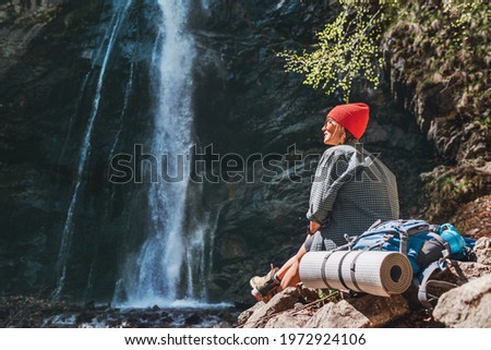 Smiling Woman with backpack dressed in active trekking clothes sitting near the mountain river waterfall and enjoying the splashing Nature power. Traveling, trekking and nature concept image.