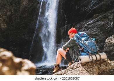 Smiling woman with backpack in red hat dressed in active trekking clothes and boots sitting near mountain river waterfall and enjoying splashing Nature power. Traveling, trekking, nature concept image - Powered by Shutterstock